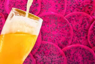 Um copo sendo preenchido com cerveja dourada, enquanto fatias de pitaya rosa compõem o fundo da imagem.