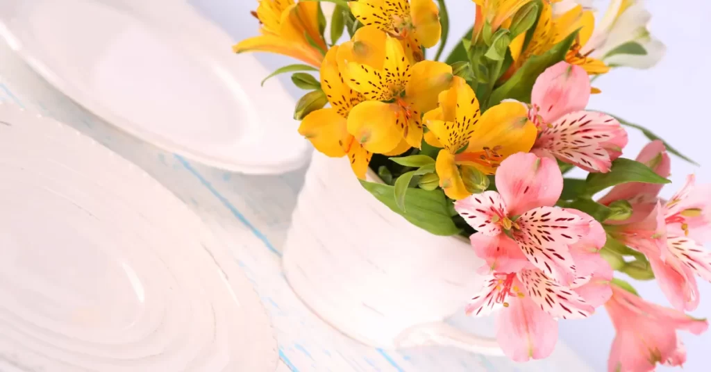 Mesa para o Dia das Mães decorada com arranjo de flores amarelas e rosas em vaso branco, ao lado de pratos brancos.