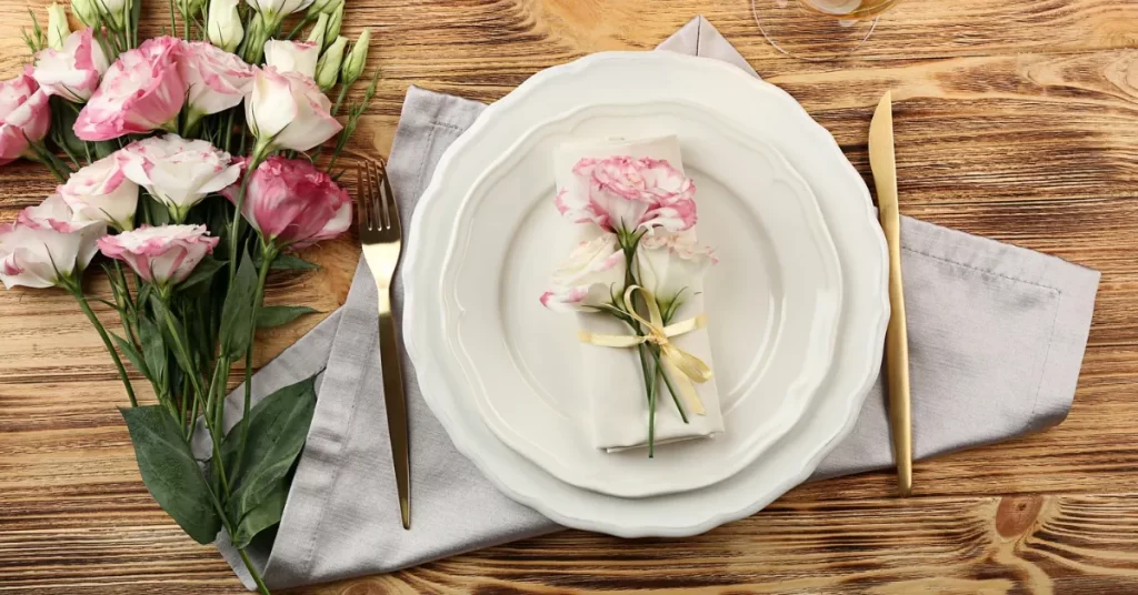 Mesa para o Dia das Mães com pratos brancos sobre guardanapo cinza, talheres dourados e arranjo de flores cor-de-rosa, incluindo flor sobre o guardanapo amarrado com fita.