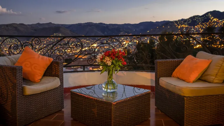 Varanda mais charmosa com poltronas de vime, almofadas laranja, mesa de centro com vaso de flores e vista panorâmica para a cidade iluminada ao entardecer.